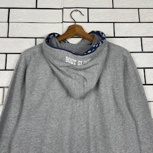 K&ouml;nnte beinhalten: Ein grauer Kapuzenpullover mit dem Text "BODY GLOVE" auf der Kapuze. Das Innere der Kapuze hat ein gemustertes Design. Der Pullover h&auml;ngt an einem Holzkleiderb&uuml;gel vor einer wei&szlig;en Backsteinmauer.
