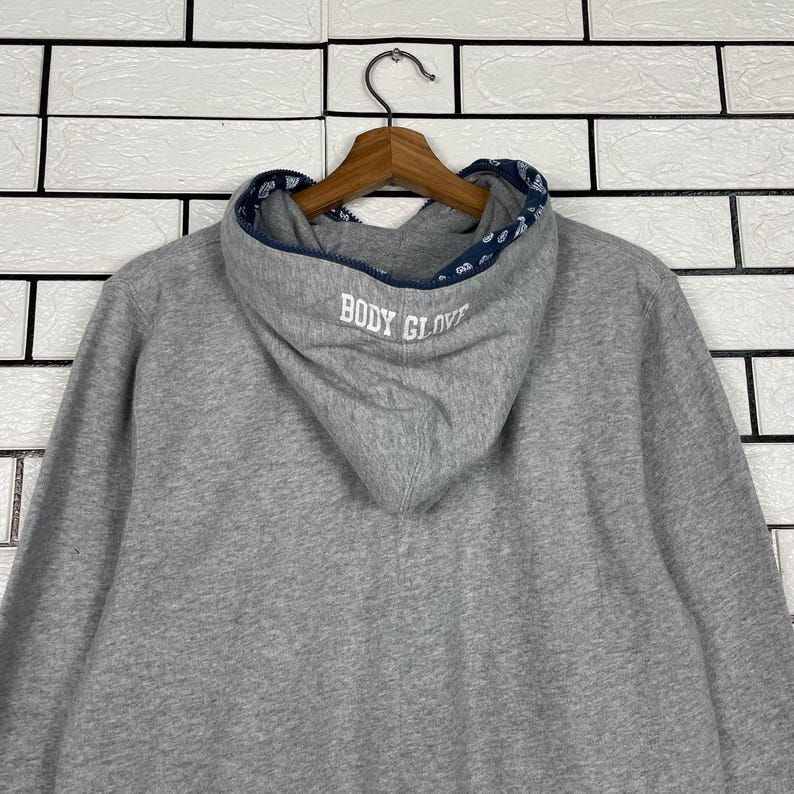 K&ouml;nnte beinhalten: Ein grauer Kapuzenpullover mit dem Text "BODY GLOVE" auf der Kapuze. Das Innere der Kapuze hat ein gemustertes Design. Der Pullover h&auml;ngt an einem Holzkleiderb&uuml;gel vor einer wei&szlig;en Backsteinmauer.
