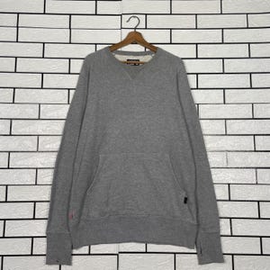 May include: A heather gray sweatshirt with a V-neck and a kangaroo pocket. The long-sleeved top has a small red accent on the left sleeve and a black tag on the right side. The sweatshirt is hanging on a wooden hanger.