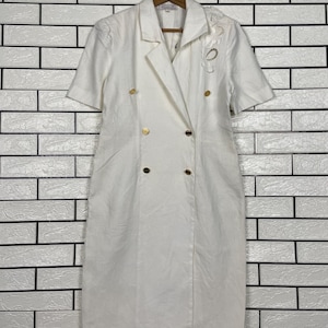 May include: White short-sleeved dress with a double-breasted design and gold-colored buttons. The dress has a notched collar and a decorative floral appliqué on the right shoulder. The dress is hanging on a wooden hanger.