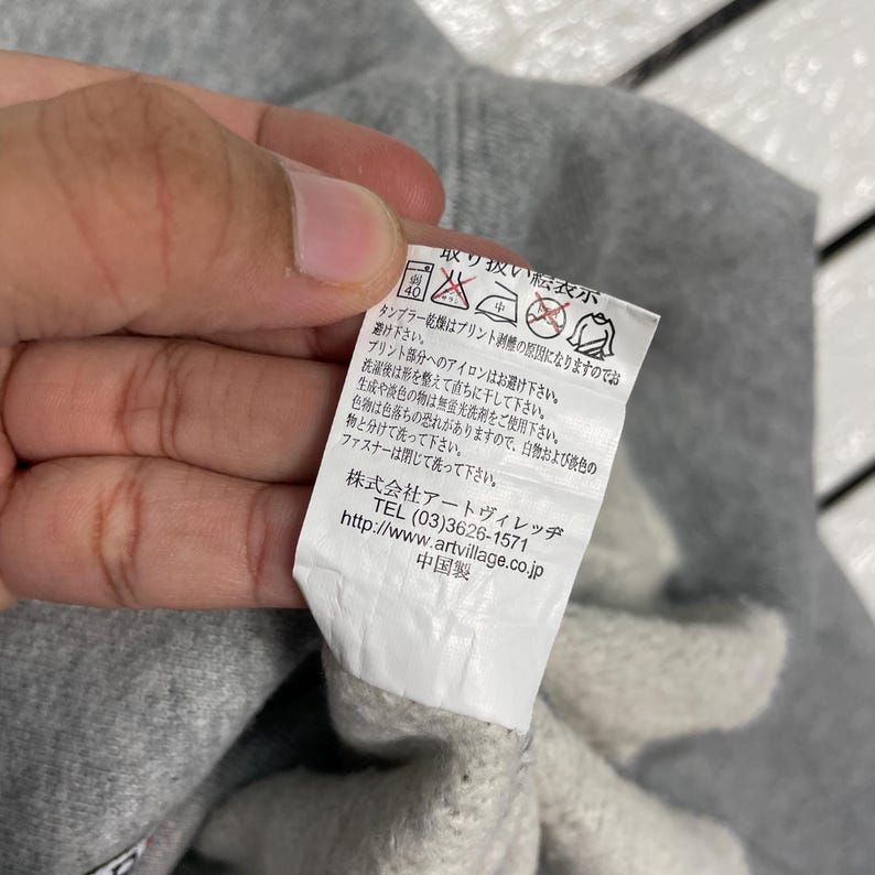K&ouml;nnte beinhalten: Nahaufnahme eines wei&szlig;en Pflegeetiketts f&uuml;r Kleidung mit japanischem Text und Pflegesymbolen. Das Etikett wird von einer Person gehalten. Das Etikett enth&auml;lt Waschanweisungen und Kontaktinformationen.