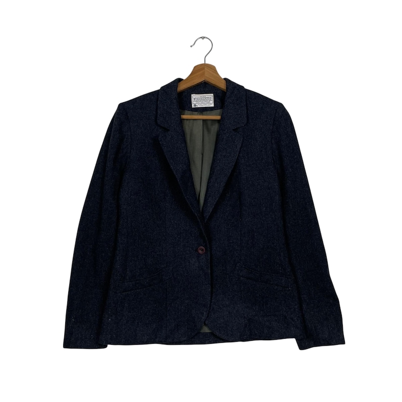 May include: A dark blue blazer with a single-button fastening, suspended from a wooden hanger. The blazer features a traditional notched lapel and a slightly textured finish. The interior lining is a muted green.