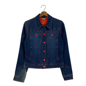 May include: A dark blue denim jacket with orange stitching and bright orange buttons. The jacket has a classic collar and a fitted silhouette. The interior lining is orange. The jacket is hanging on a wooden hanger.