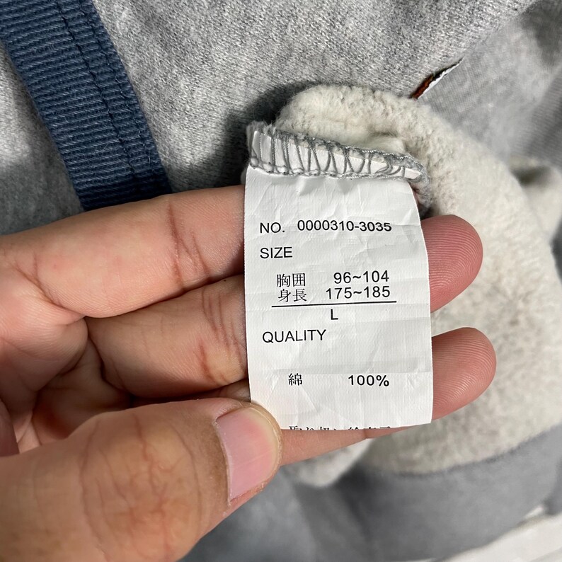 K&ouml;nnte beinhalten: Nahaufnahme eines grauen Sweatshirt-Etiketts mit dem Text "NO. 0000310-3035", "SIZE" und japanischen Schriftzeichen. Das Etikett zeigt auch Brustumfang 96-104 und K&ouml;rpergr&ouml;&szlig;e 175-185. Das Material ist 100% Baumwolle.