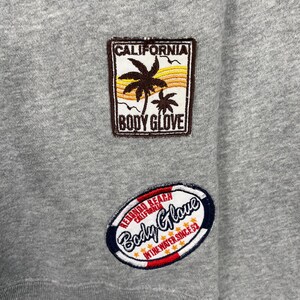 K&ouml;nnte beinhalten: Nahaufnahme eines grauen Sweatshirts mit zwei gestickten Aufn&auml;hern. Der obere Aufn&auml;her zeigt "CALIFORNIA" &uuml;ber einer Palmen-Grafik und "BODY GLOVE". Der untere Aufn&auml;her zeigt "REDONDO BEACH CALIFORNIA" und "Body Glove" mit "IN THE WATER SINCE 53."