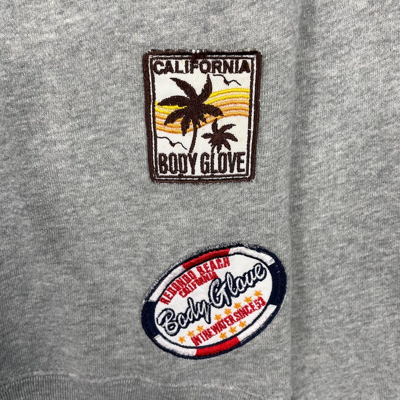 K&ouml;nnte beinhalten: Nahaufnahme eines grauen Sweatshirts mit zwei gestickten Aufn&auml;hern. Der obere Aufn&auml;her zeigt "CALIFORNIA" &uuml;ber einer Palmen-Grafik und "BODY GLOVE". Der untere Aufn&auml;her zeigt "REDONDO BEACH CALIFORNIA" und "Body Glove" mit "IN THE WATER SINCE 53."