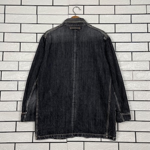 May include: A dark grey denim jacket with long sleeves, a collar, and button closure. The jacket has a faded appearance and is hanging on a metal hanger against a white brick wall. The jacket appears to be a medium length.