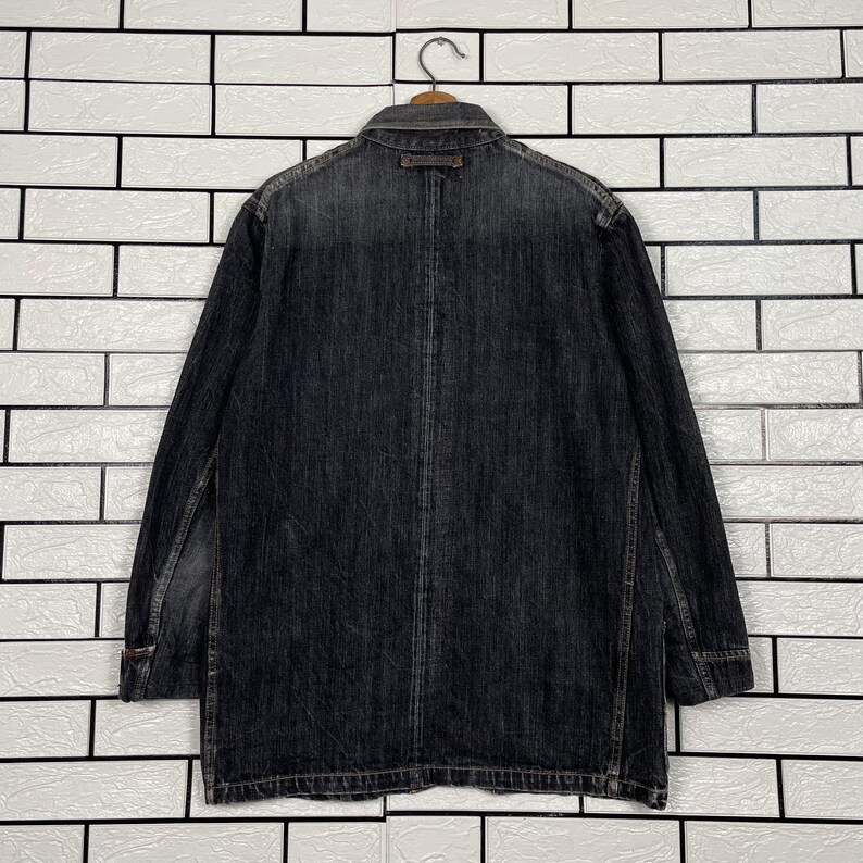 May include: A dark grey denim jacket with long sleeves, a collar, and button closure. The jacket has a faded appearance and is hanging on a metal hanger against a white brick wall. The jacket appears to be a medium length.