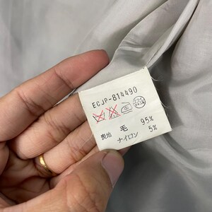 May include: A close-up of a label with the text "ECJP-814490" and fabric care symbols. The label also indicates the fabric composition: 95% wool and 5% nylon. The background is a light grey fabric.