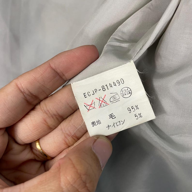 May include: A close-up of a label with the text "ECJP-814490" and fabric care symbols. The label also indicates the fabric composition: 95% wool and 5% nylon. The background is a light grey fabric.