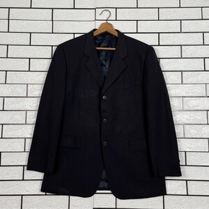 以下が含まれることがあります： ノッチドラペル、3つボタンのフロントクロージャー、ウエストに2つのフラップポケットを備えた無地の黒いブレザー。木製のハンガーに掛けられており、長袖とダークな裏地が付いた構造的なシルエットが特徴です。