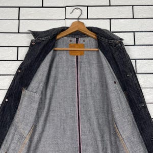 May include: A dark grey denim jacket with a classic collar and button fastenings. The jacket is suspended on a wooden hanger. The interior lining is grey with a pink stripe down the centre. A brown tag with the number "505" is visible.
