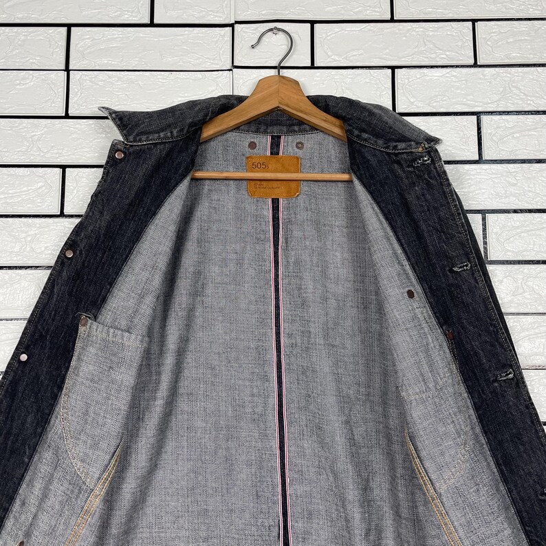 May include: A dark grey denim jacket with a classic collar and button fastenings. The jacket is suspended on a wooden hanger. The interior lining is grey with a pink stripe down the centre. A brown tag with the number "505" is visible.