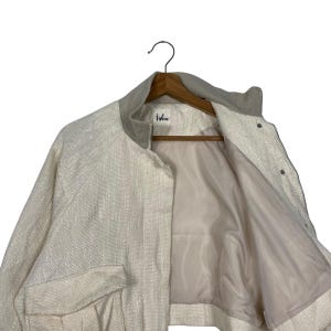 May include: Off-white jacket with a textured weave, featuring a round collar and two front pockets. The jacket has a zipper closure and snap buttons. The interior lining is a light beige color. The brand name "igloo" is visible on the label.
