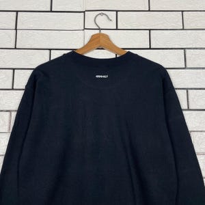 May include: A black sweatshirt with the word "GRAMICCI" printed in white on the upper back. The sweatshirt is hanging on a wooden hanger against a white brick wall.