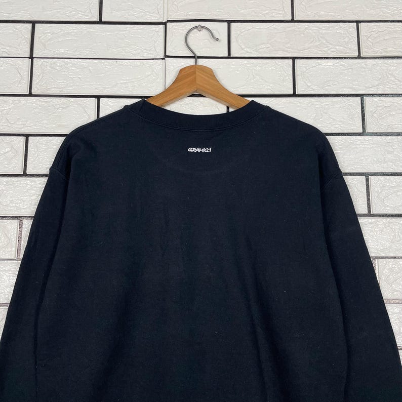 May include: A black sweatshirt with the word "GRAMICCI" printed in white on the upper back. The sweatshirt is hanging on a wooden hanger against a white brick wall.
