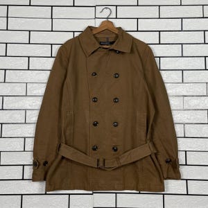 以下が含まれることがあります： 茶色のダブルブレストコートと、お揃いのベルト。ノッチカラーと複数のダークボタンを備えたクラシックなデザインです。白いレンガの壁を背景に、木製のハンガーにコートが掛かっています。