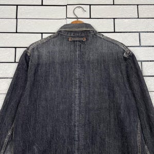 May include: A dark grey denim jacket, seen from the back, suspended on a wooden hanger. The jacket features a collar, button details, and a worn look. The backdrop is a white brick wall.