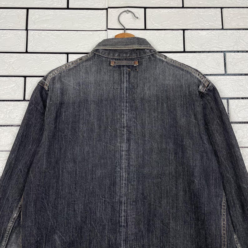 May include: A dark grey denim jacket, seen from the back, suspended on a wooden hanger. The jacket features a collar, button details, and a worn look. The backdrop is a white brick wall.