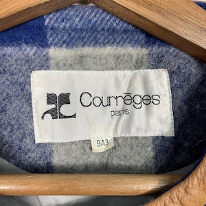 May include: A close-up of a Courr&egrave;ges Paris label on a jacket. The label is white with black text and a logo. The jacket has a blue and grey plaid pattern.