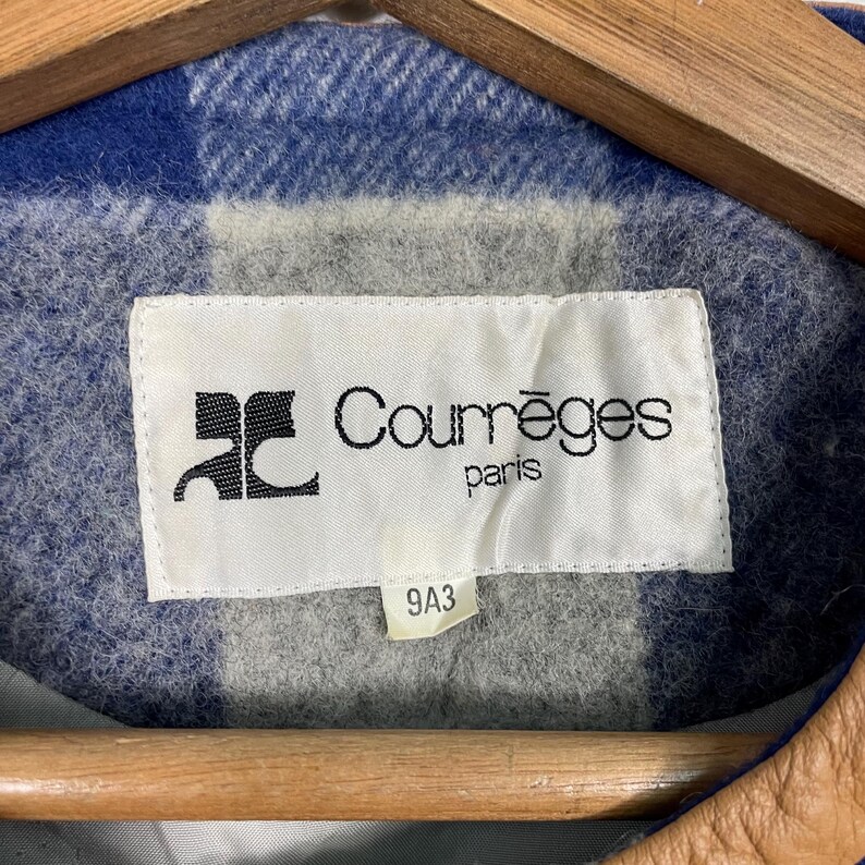May include: A close-up of a Courr&egrave;ges Paris label on a jacket. The label is white with black text and a logo. The jacket has a blue and grey plaid pattern.