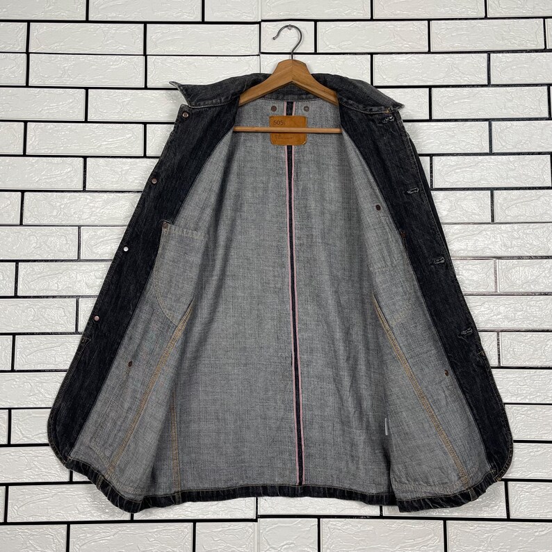 May include: A dark grey denim jacket hanging on a wooden hanger. The jacket is open, revealing a grey interior lining with a pink stripe down the centre. The jacket has two front pockets and button closures.