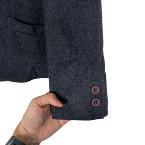 May include: Close-up of a dark grey blazer sleeve with two brown buttons. The blazer has a pocket and is made of a textured fabric. The sleeve is slightly angled, showing the cuff and buttons.