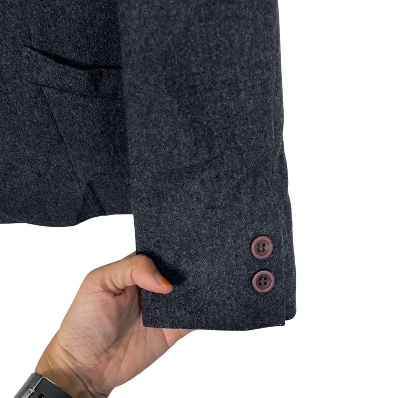 May include: Close-up of a dark grey blazer sleeve with two brown buttons. The blazer has a pocket and is made of a textured fabric. The sleeve is slightly angled, showing the cuff and buttons.