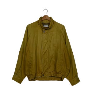 May include: Olive green jacket with a stand-up collar and a button closure. The jacket has long sleeves, two front pockets, and a ribbed hem. The brand name "OGREY" is visible on the collar. The jacket is hanging on a wooden hanger.