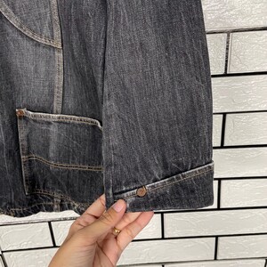 May include: Close-up of a dark grey denim jacket. The jacket has a buttoned cuff, a front pocket, and visible stitching. The fabric has a textured appearance, and the jacket appears to be well-worn.