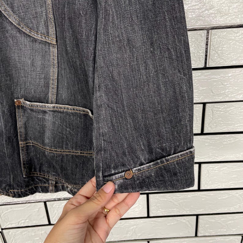 May include: Close-up of a dark grey denim jacket. The jacket has a buttoned cuff, a front pocket, and visible stitching. The fabric has a textured appearance, and the jacket appears to be well-worn.