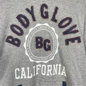 K&ouml;nnte beinhalten: Nahaufnahme eines grauen Sweatshirts mit den Worten "BODY GLOVE" in einer Schriftart im Denim-Stil. Das Sweatshirt hat auch ein kreisf&ouml;rmiges Emblem mit "BG" und den Worten "CALIFORNIA SURF GROOVE". Das Wort "CALIFORNIA" ist in gro&szlig;en wei&szlig;en Buchstaben gedruckt.