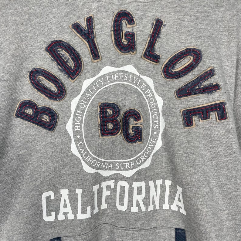 K&ouml;nnte beinhalten: Nahaufnahme eines grauen Sweatshirts mit den Worten "BODY GLOVE" in einer Schriftart im Denim-Stil. Das Sweatshirt hat auch ein kreisf&ouml;rmiges Emblem mit "BG" und den Worten "CALIFORNIA SURF GROOVE". Das Wort "CALIFORNIA" ist in gro&szlig;en wei&szlig;en Buchstaben gedruckt.