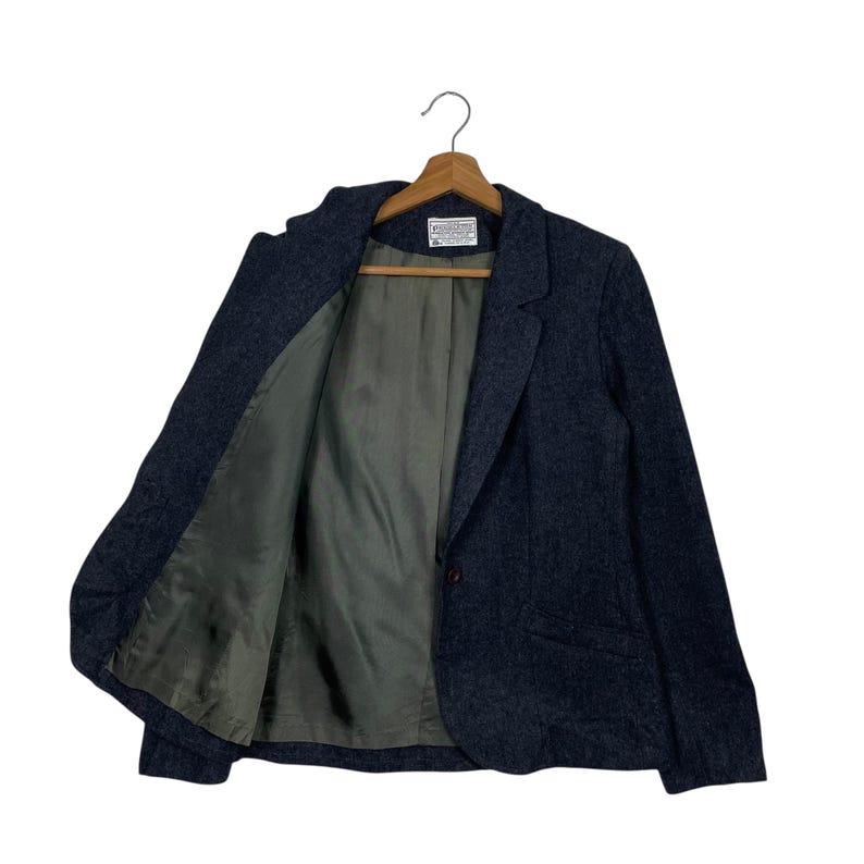 May include: A dark blue blazer with a single-button fastening, suspended from a wooden hanger. The blazer has a textured finish and a green lining. The jacket is open, showing the lining and the label.