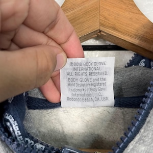 K&ouml;nnte beinhalten: Nahaufnahme eines grauen Hoodie-Etiketts mit Text. Auf dem Etikett steht "&copy;2010 BODY GLOVE INTERNATIONAL. ALLE RECHTE VORBEHALTEN. BODY GLOVE und das Hand Design sind eingetragene Marken von Body Glove International, LLC, Redondo Beach, CA. USA."