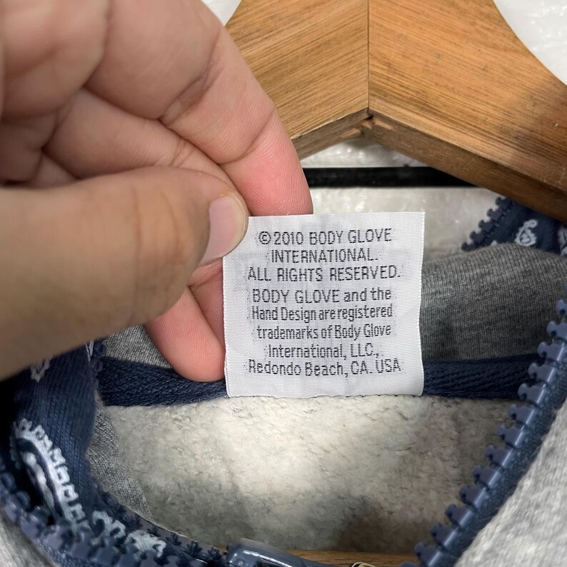 K&ouml;nnte beinhalten: Nahaufnahme eines grauen Hoodie-Etiketts mit Text. Auf dem Etikett steht "&copy;2010 BODY GLOVE INTERNATIONAL. ALLE RECHTE VORBEHALTEN. BODY GLOVE und das Hand Design sind eingetragene Marken von Body Glove International, LLC, Redondo Beach, CA. USA."