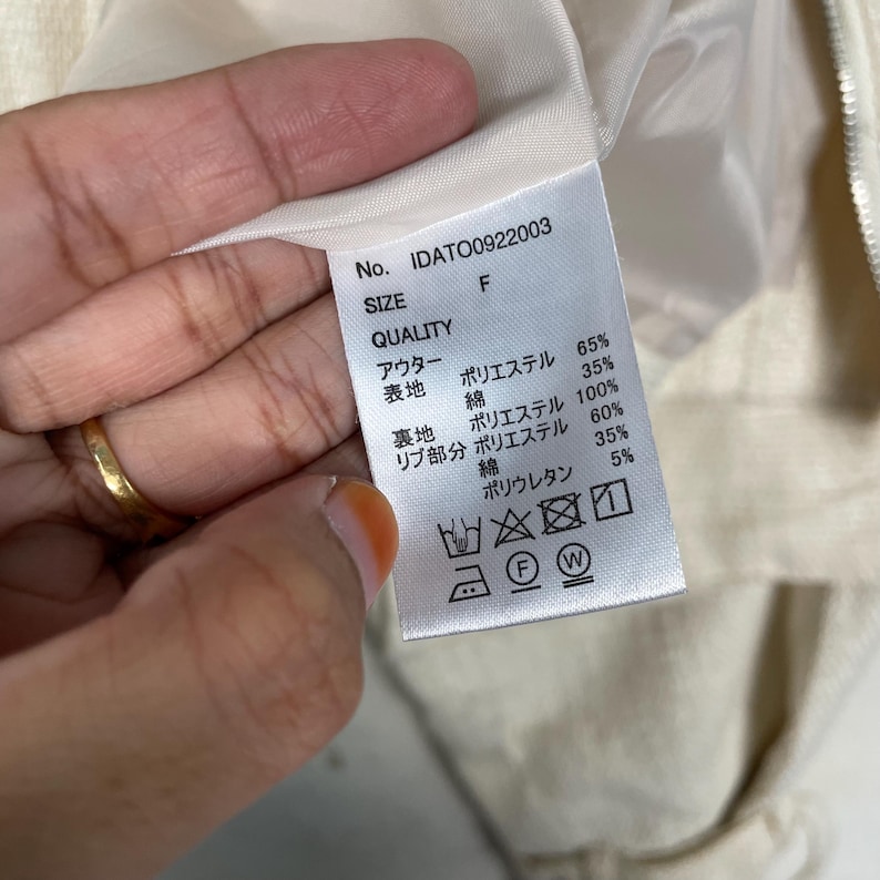 May include: A close-up of a tag with text in Japanese and English. The tag includes the number IDATO0922003, size F, and fabric composition percentages. The tag is attached to a light-colored garment.