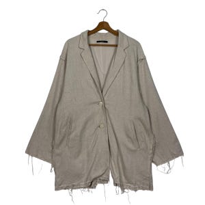 Könnte beinhalten: Beiger Blazer mit einem Knopfverschluss und ausgefransten Kanten. Die langärmlige Jacke hat ein fallendes Revers und zwei Vordertaschen. Das Kleidungsstück hängt an einem Holzbügel.