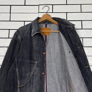 May include: A dark grey denim jacket with a button-up front and a collared neckline. The jacket is hanging on a wooden hanger. The interior lining is grey with a pink accent. A brown tag is visible.