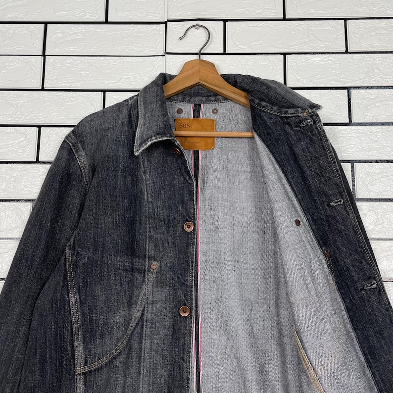 May include: A dark grey denim jacket with a button-up front and a collared neckline. The jacket is hanging on a wooden hanger. The interior lining is grey with a pink accent. A brown tag is visible.
