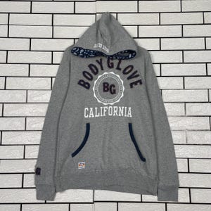 K&ouml;nnte beinhalten: Grauer Kapuzenpullover mit dem Text "BODY GLOVE CALIFORNIA" in Wei&szlig; und Marineblau. Der Pullover hat eine K&auml;ngurutasche und eine marineblau gef&uuml;tterte Kapuze. Das Body Glove-Logo ist mittig auf der Brust platziert.