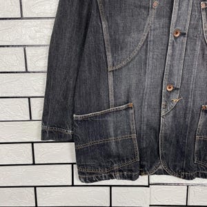 May include: A dark grey denim jacket with a tailored fit. The jacket has a button-up front, two front pockets, and a notched lapel. The stitching is brown, and the jacket has a vintage appearance.