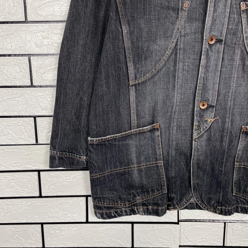 May include: A dark grey denim jacket with a tailored fit. The jacket has a button-up front, two front pockets, and a notched lapel. The stitching is brown, and the jacket has a vintage appearance.