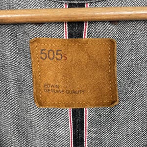 May include: Close-up of a denim jacket with a brown leather patch. The patch reads "505s" and "EDWIN GENUINE QUALITY" in a serif font. The jacket is grey with red and white stitching.