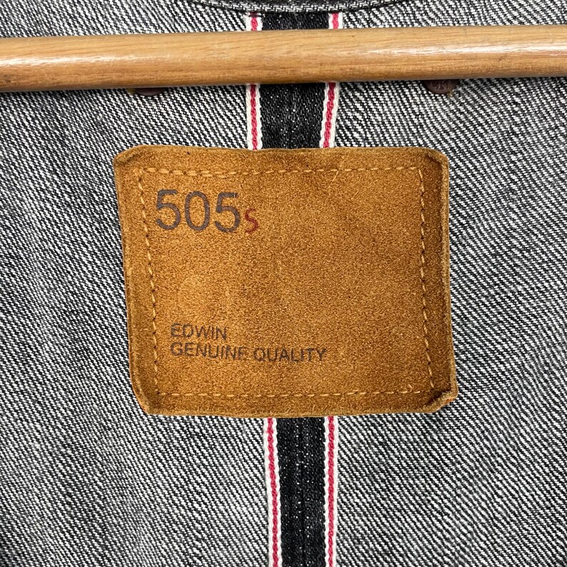May include: Close-up of a denim jacket with a brown leather patch. The patch reads "505s" and "EDWIN GENUINE QUALITY" in a serif font. The jacket is grey with red and white stitching.