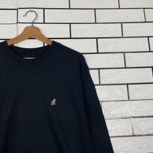 May include: Black crew neck sweatshirt with a small white embroidered logo on the chest. The sweatshirt is hanging on a wooden hanger against a white brick wall.