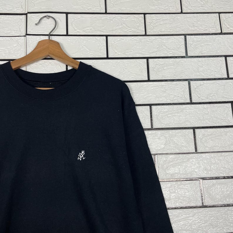 May include: Black crew neck sweatshirt with a small white embroidered logo on the chest. The sweatshirt is hanging on a wooden hanger against a white brick wall.