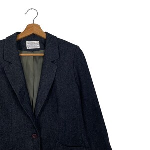 May include: A dark grey blazer with a classic design, hanging on a wooden hanger. The blazer features a notched lapel, a single-button closure, and a label on the inside. The lining is a muted green colour.