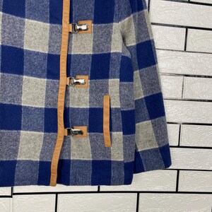May include: A blue, grey, and white checked jacket featuring brown leather detailing and silver metal toggle fastenings. The jacket has a traditional design, a front pocket, and a warm, textured appearance.