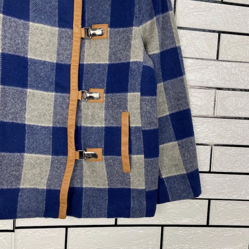 May include: A blue, grey, and white checked jacket featuring brown leather detailing and silver metal toggle fastenings. The jacket has a traditional design, a front pocket, and a warm, textured appearance.